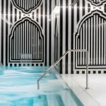 Zona de aguas spa con piscina y cascadas de de agua se ve la piscina de color azul el suelo de mármol y ñas paredes vas de rayas negras y blancas con arcos nazari